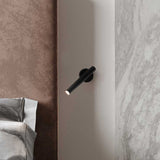 Rui LED Adjustable Wall Light with Reading Light in matt black, slim cylindrical tube on a short pivoting arm, mounted bedside above a bed with soft fabric headboard and marble wall panel.