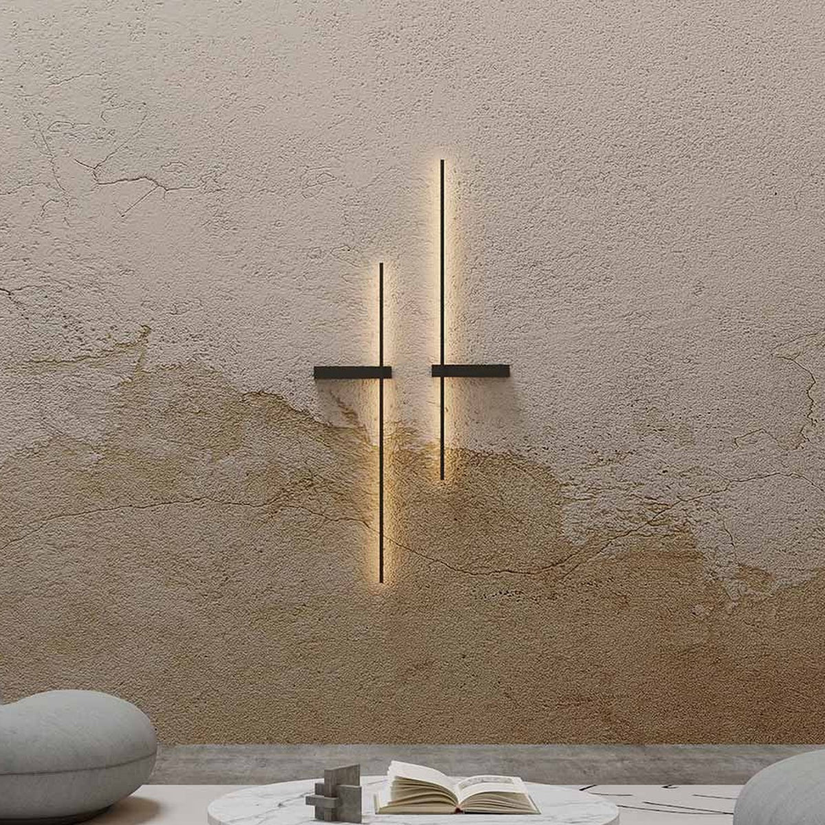 Rut LED Linear Wall Light in matt black, two vertical bars with horizontal mounting brackets casting warm uplighting on a textured plaster wall in a living room setting.