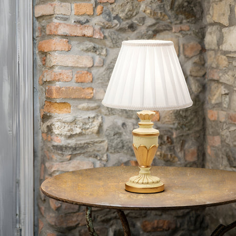 The Firenze Table Lamp - White, a vintage gem featuring a pleated white shade, adorns a round wooden table. Its cream and gold base embodies classic design allure set against a rustic stone brick wall, making it a versatile choice for any setting.