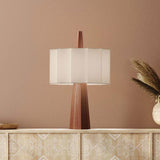Rain Scalloped Fabric Table Lamp in chestnut, tapered faceted wood base with a central spire and white scalloped fabric shade, shown on a sideboard against a pink wall.