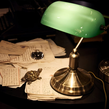 A vintage desk with the Trellis Table Lamp in brass, showcasing a green glass diffuser, illuminates a stack of handwritten letters. A small decorative turtle figurine and a round glass paperweight rest on the papers, creating a nostalgic, classic ambiance.