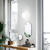 A minimalistic vanity setup by a window showcases two oval mirrors, a small circular mirror, and assorted makeup items. The area is illuminated by the Orlix Ceiling & Wall Light in white blown glass, measuring 14cm, while potted plants add a touch of greenery to the compact space.