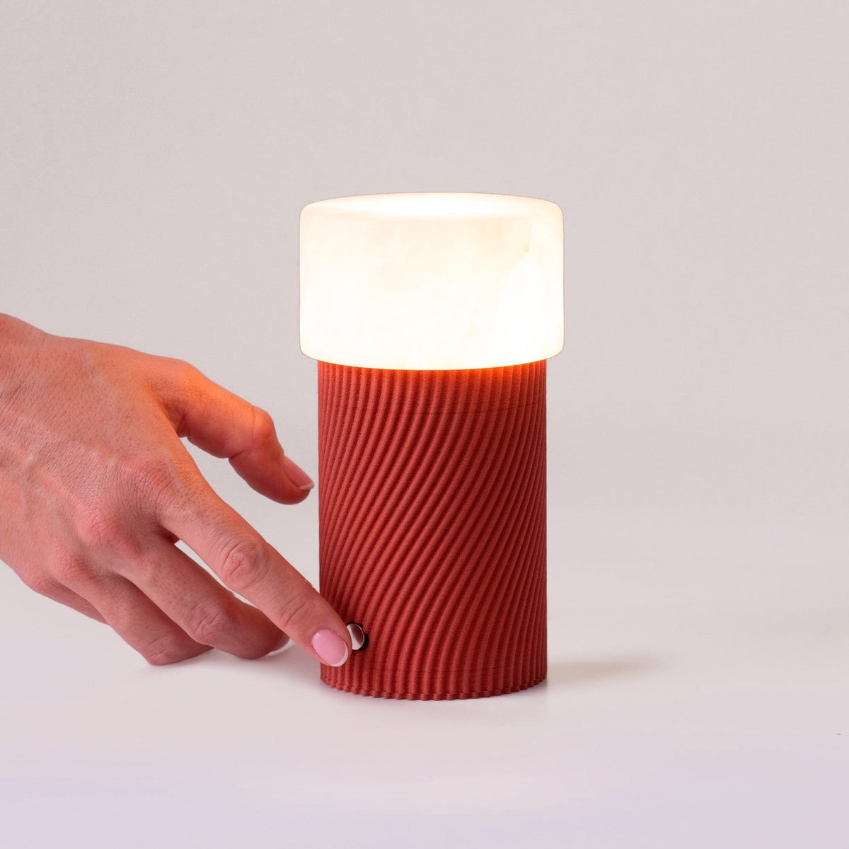Rizzo LED alabaster rechargeable table lamp with a ribbed Sapelly red cylindrical base and white alabaster shade, shown glowing as a hand touches its base control on a plain background.