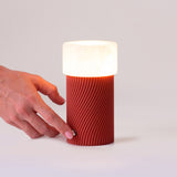 Rizzo LED alabaster rechargeable table lamp with a ribbed Sapelly red cylindrical base and white alabaster shade, shown glowing as a hand touches its base control on a plain background.
