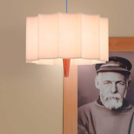 Rain Scalloped Fabric Pendant Light in Chestnut, white fabric shade with scalloped edges, tapered wood centre, blue textile cable, shown near a framed portrait.