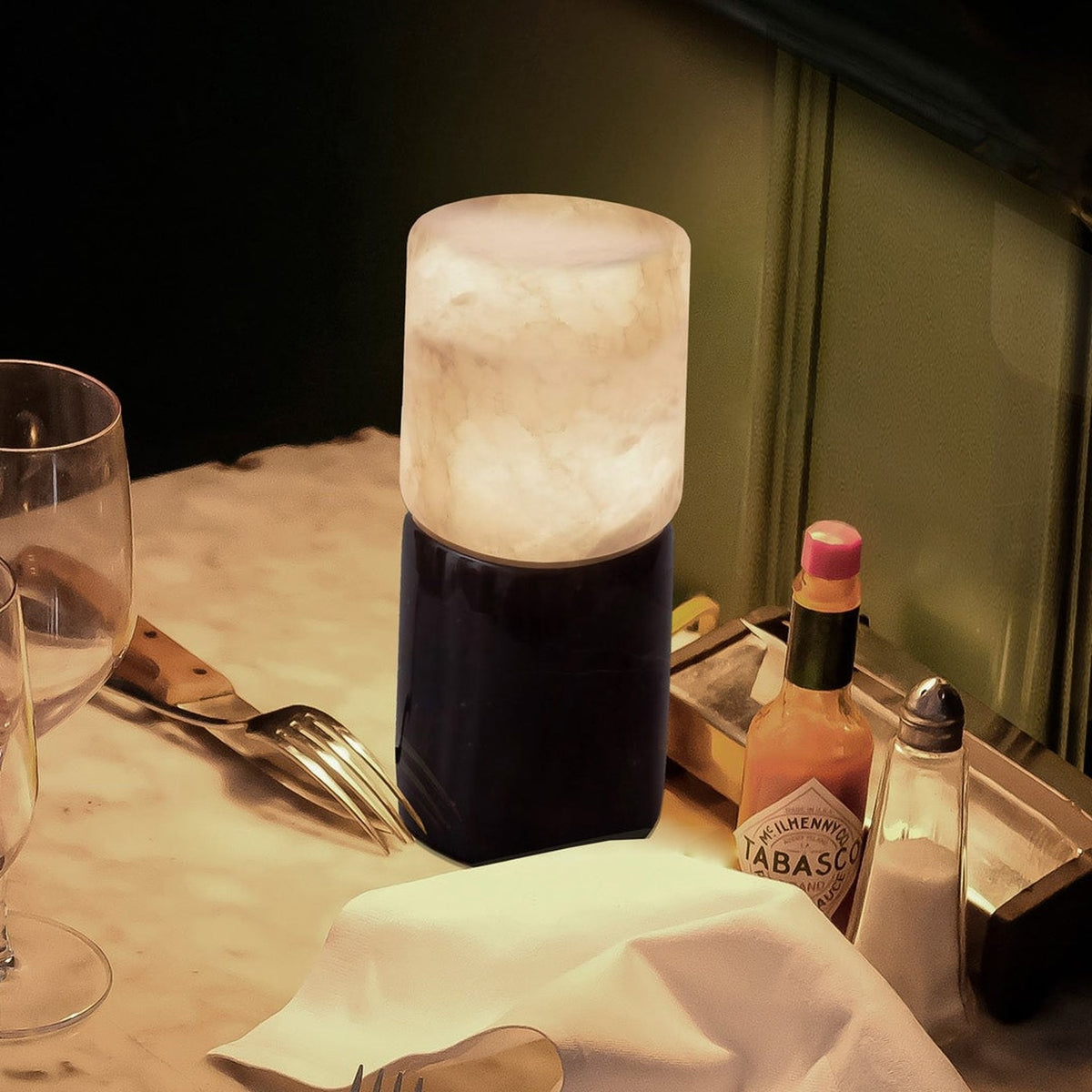 Rock Cylinder Alabaster Table Lamp with dark marble base, lit on a restaurant table beside glassware, cutlery, and a Tabasco bottle.