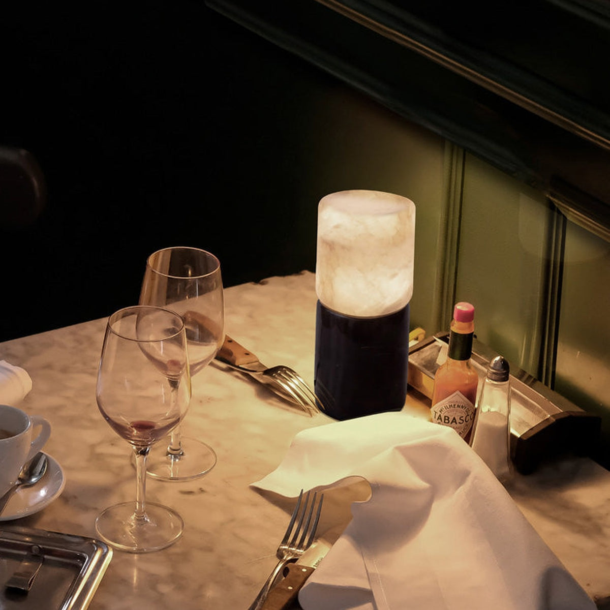 Rock Cylinder Alabaster Table Lamp with dark marble base, lit on a marble restaurant table beside wine glasses, cutlery, and a napkin.