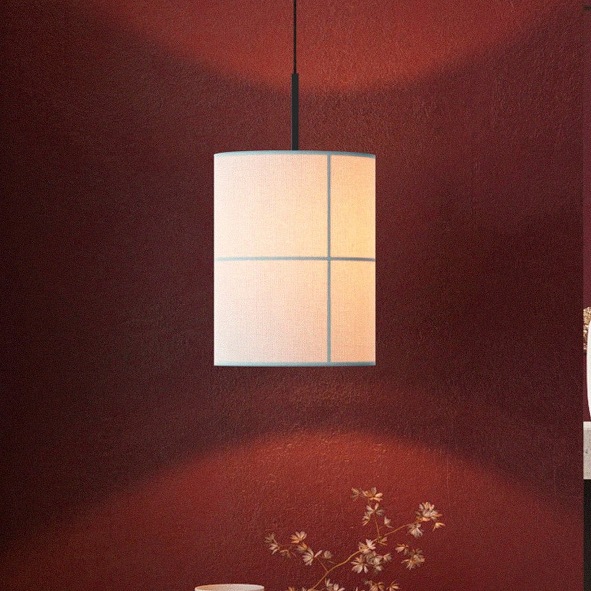 Rania cylinder pendant light with white cream fabric shade and fine grid seams, hung from a matt black iron fitting against a dark red wall.