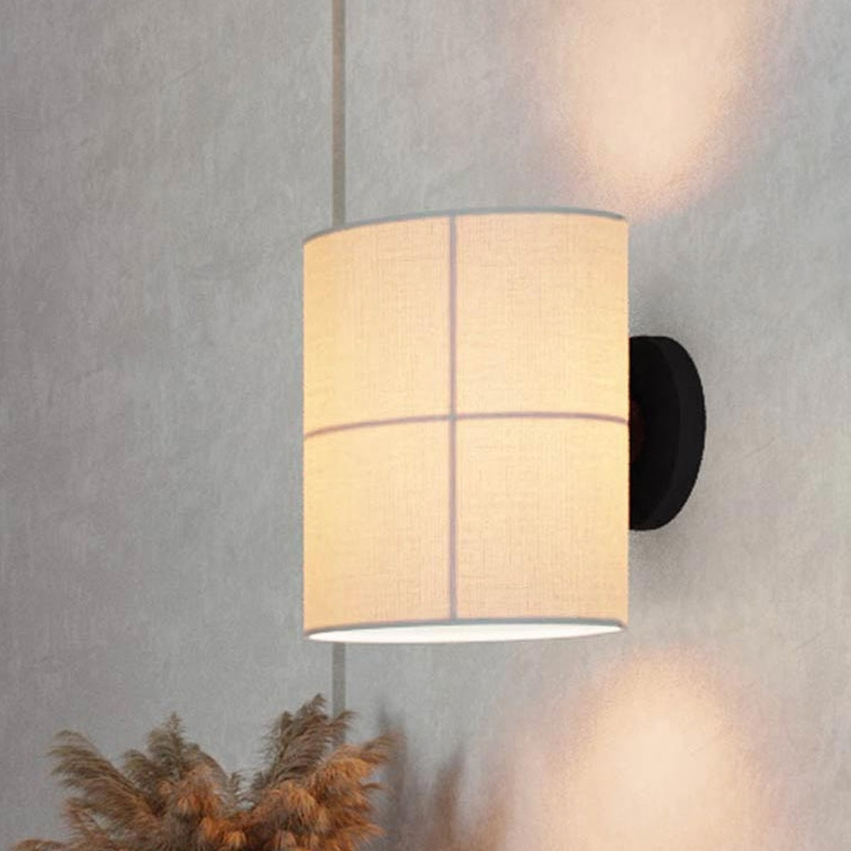 Rania Cylinder Fabric Wall Light in white cream with cross-line detailing on a round black base, mounted on a grey wall above a dried plant.
