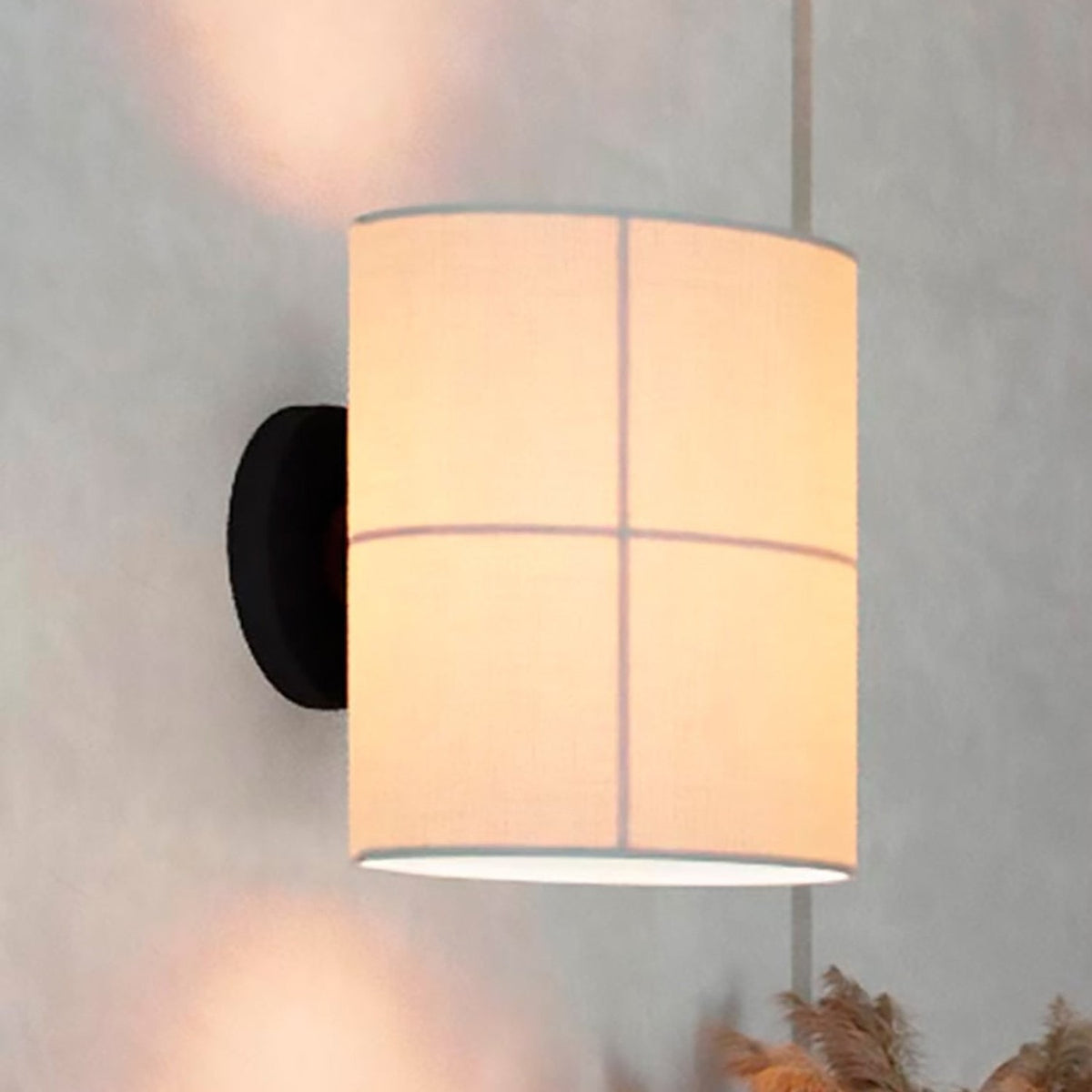 Rania cylinder fabric wall light with a white cream shade divided into four panels by fine lines, mounted on a round black base against a grey wall.