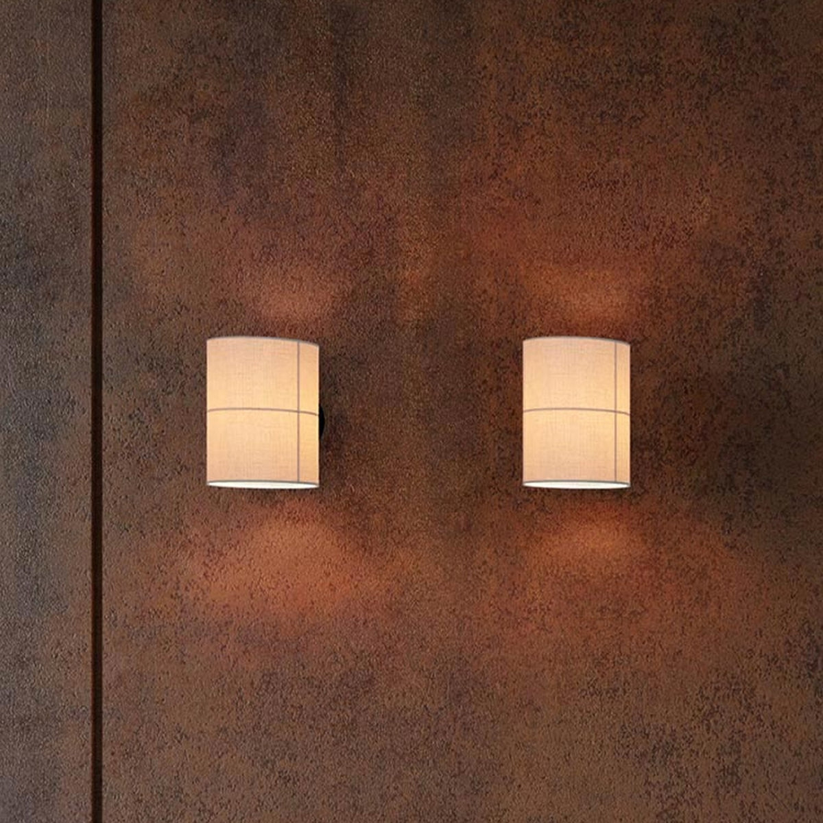 Rania Cylinder Fabric Wall Light in white cream with black base, two fitted on a textured rust-brown wall, glowing softly uplight and downlight.
