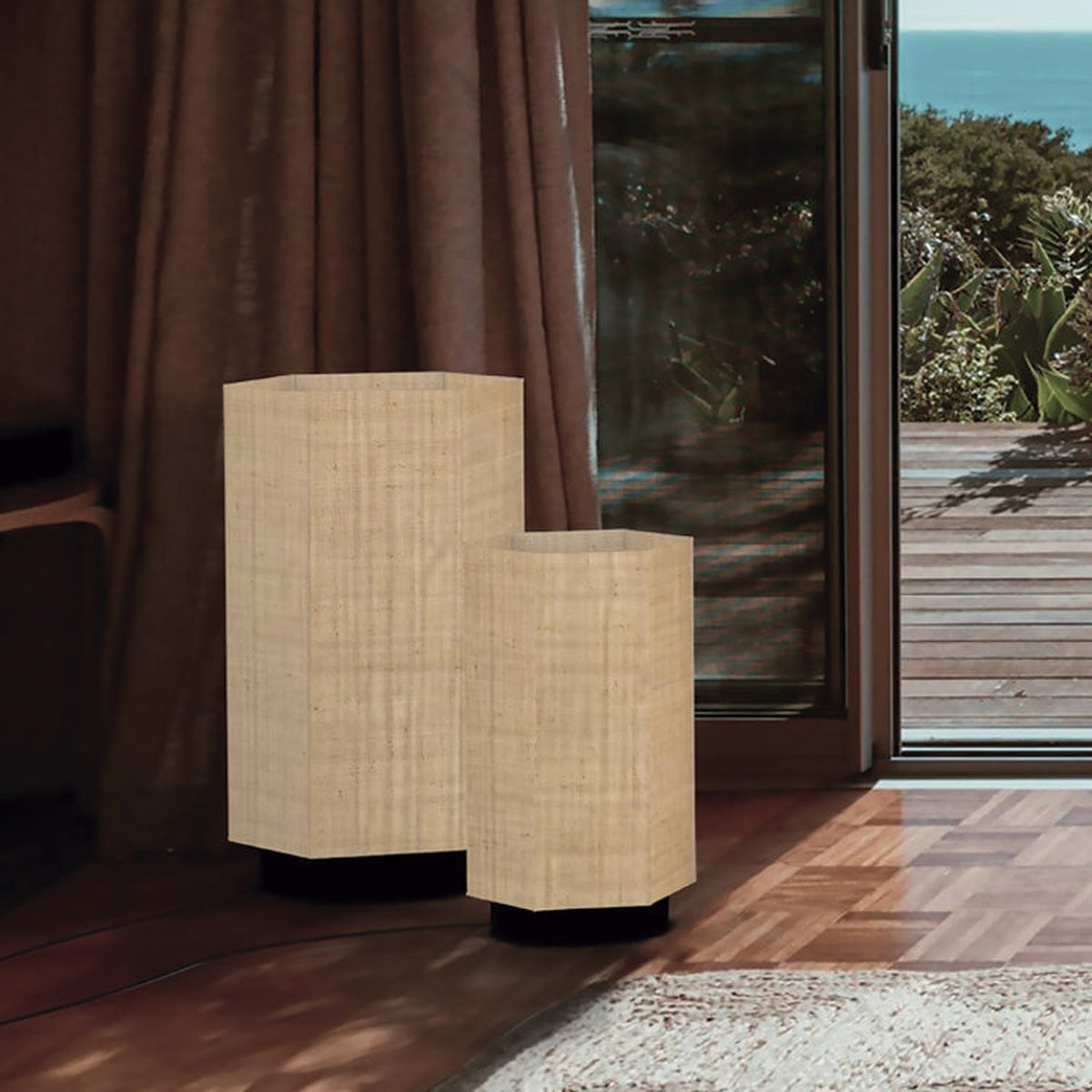 Roxi Fabric Floor Lamp in two sizes, hexagonal natural fibre shades on dark round bases, shown beside a curtained window with a sea view.