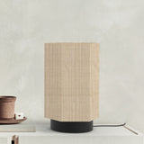 Roxi Fabric Table Lamp in natural fibre with a hexagonal woven shade and round black base, shown on a sideboard beside pottery, against a light grey wall.