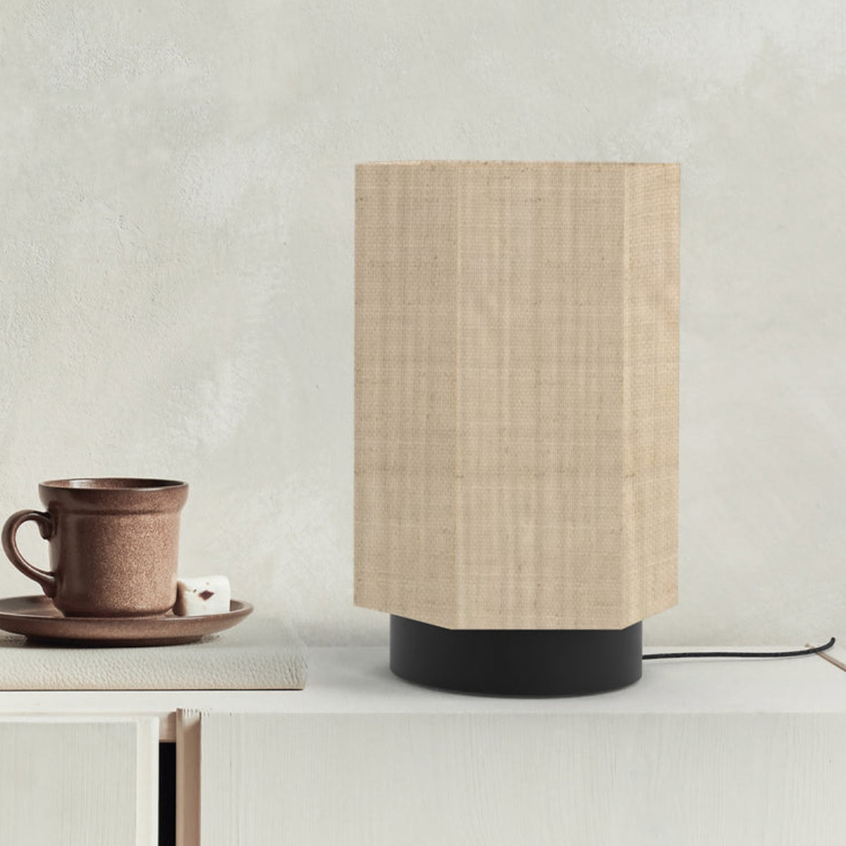Roxi Fabric Table Lamp with hexagonal natural fibre shade and round black base, shown on a sideboard beside a cup and saucer against a neutral wall.