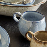 Sandrine Speckled Stoneware Mug 500ml in blue, with a rounded body, loop handle, and dark rim band, shown beside a smaller brown mug and stoneware dishes on a dark table.