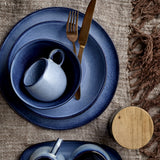 Sandrine Stoneware Cup 275ml in blue with round loop handle, set inside matching blue bowl and plates alongside gold cutlery on a woven tablecloth.