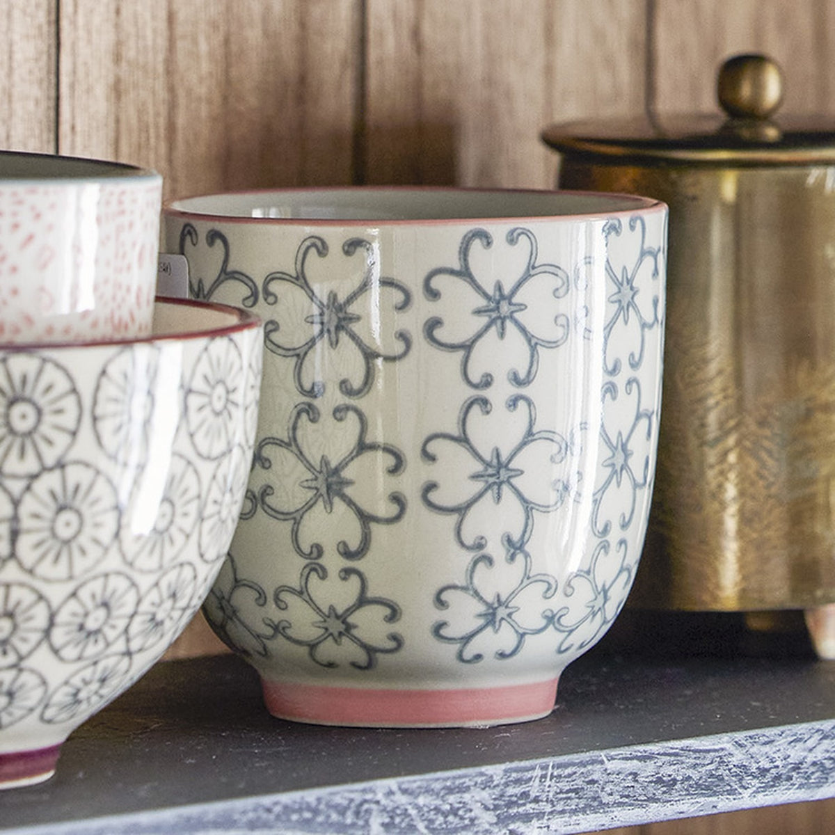 Cécile Cup 150ml in ceramic, round handleless design with grey floral decal pattern and pink rim, shown among assorted patterned cups on a shelf.