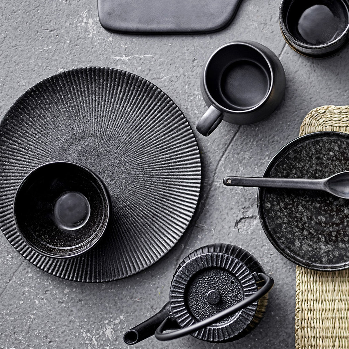 Neri Stoneware Side Plate in black with ribbed sunburst texture, styled on a grey surface alongside black stoneware cups, bowls, and a teapot.