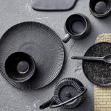 Neri Stoneware Side Plate in black with ribbed sunburst texture, styled on a grey surface alongside black stoneware cups, bowls, and a teapot.