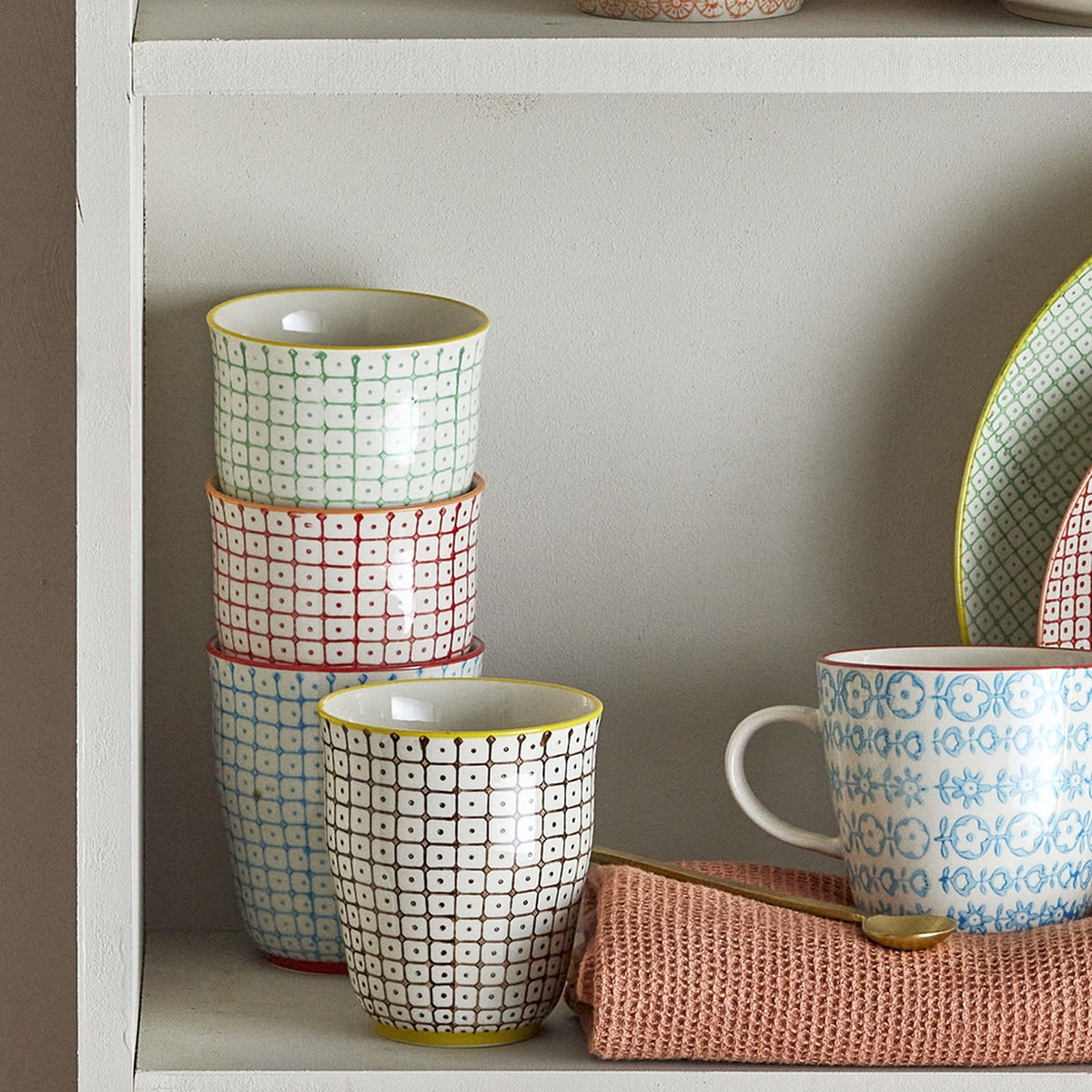 Carla handleless stoneware cups in assorted patterned designs with grid and dot motifs, stacked and displayed on a shelf alongside a patterned mug and plate.