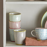Carla handleless stoneware cups in assorted patterned designs with grid and dot motifs, stacked and displayed on a shelf alongside a patterned mug and plate.