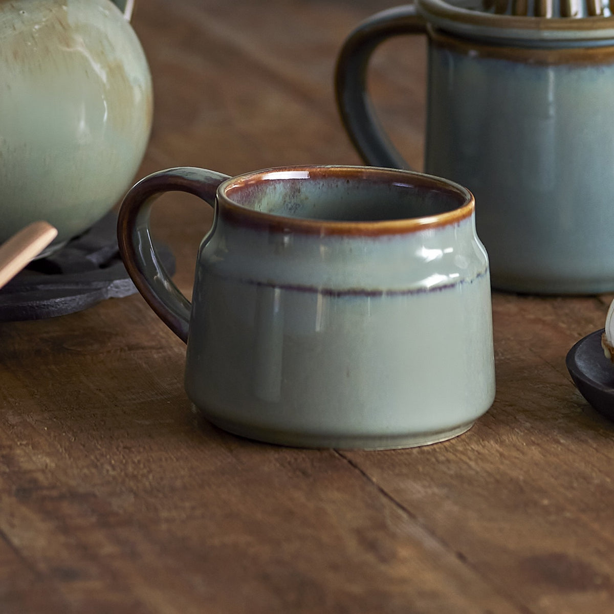 Pixie Stoneware Cup 350ml in green, wide low ceramic cup with a reactive speckled glaze and open loop handle, shown on a wooden table with teapots in the background.