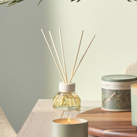 Hinoki Sage Reed Diffuser 88.7ml in a clear glass bottle with white collar and reed sticks, displayed on a table beside a lit candle and tinned candle.