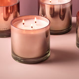 Coconut Milk Mango scented candle in a glass vessel with pink-toned finish, lit on three cotton wicks, shown alongside similar candles on a pale pink surface.
