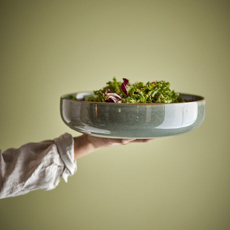Pixie Stoneware Bowl 24cm in green, shown held on an outstretched hand with a salad, showing its wide shallow shape and contrasting rim against a plain green background.