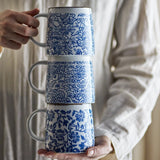 Three stacked Molly stoneware mugs with distinct blue floral patterns and white bodies, held in two hands against a person's neutral-toned shirt.