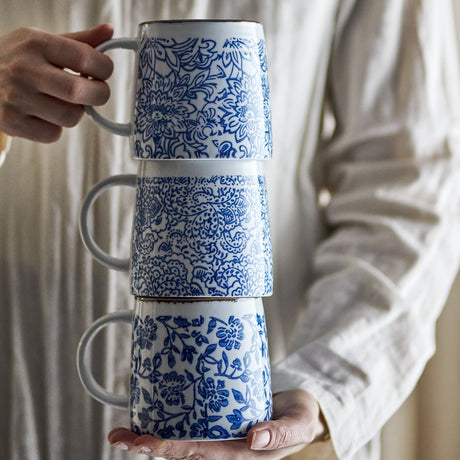 Three stacked Molly stoneware mugs with distinct blue floral patterns and white bodies, held in two hands against a person's neutral-toned shirt.