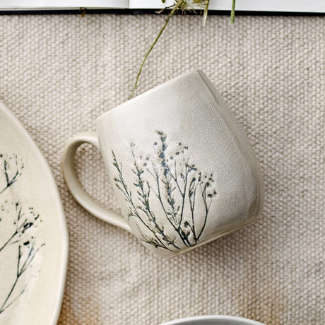 Bea Crackle Stoneware Mug 400ml in cream with crackle-glazed finish and botanical stem print, curved handle, shown beside a matching plate on a woven beige surface.