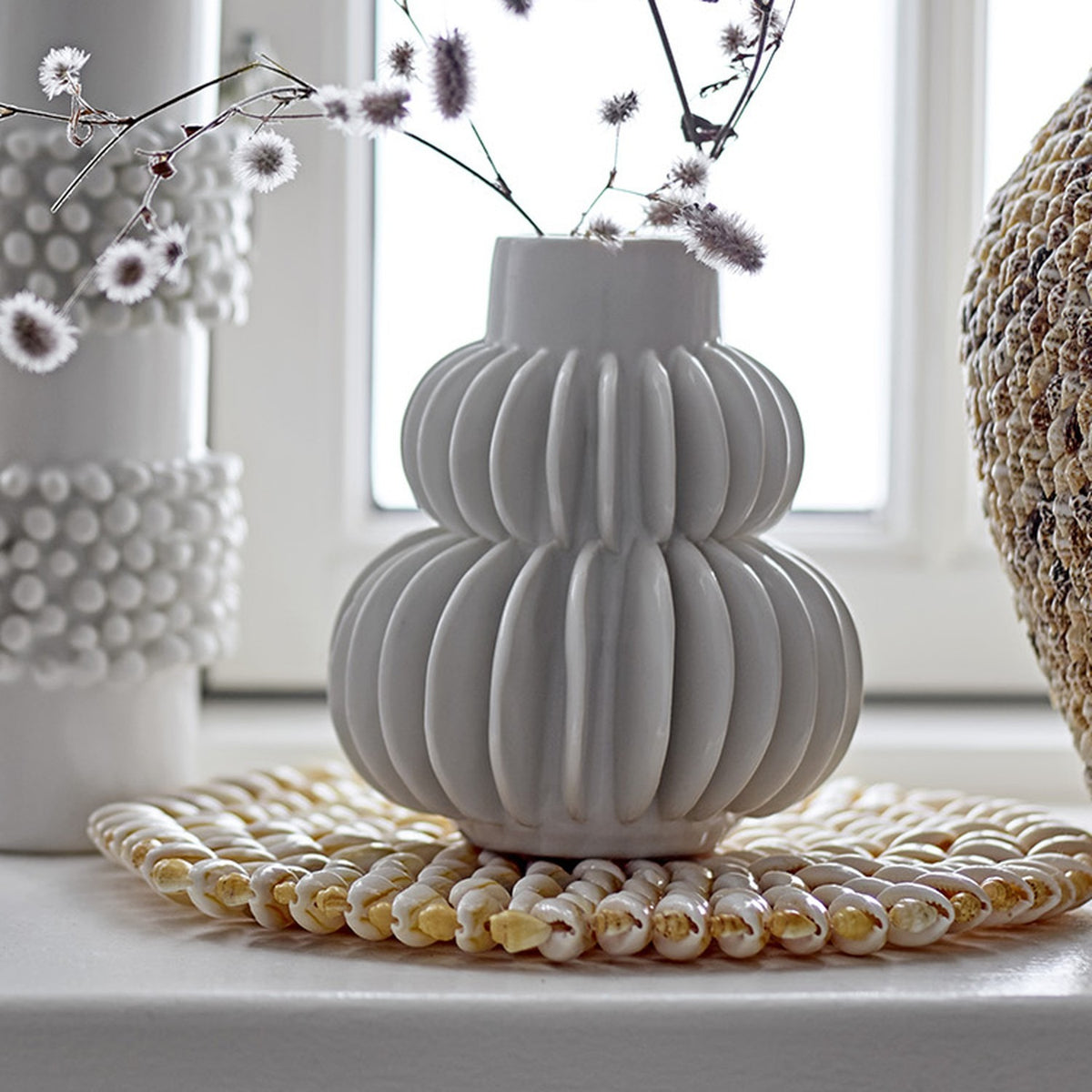 Halfdan Stoneware Vase 14cm in white with ribbed, tiered body and narrow neck, holding dried flowers on a windowsill beside textured hobnail vases.