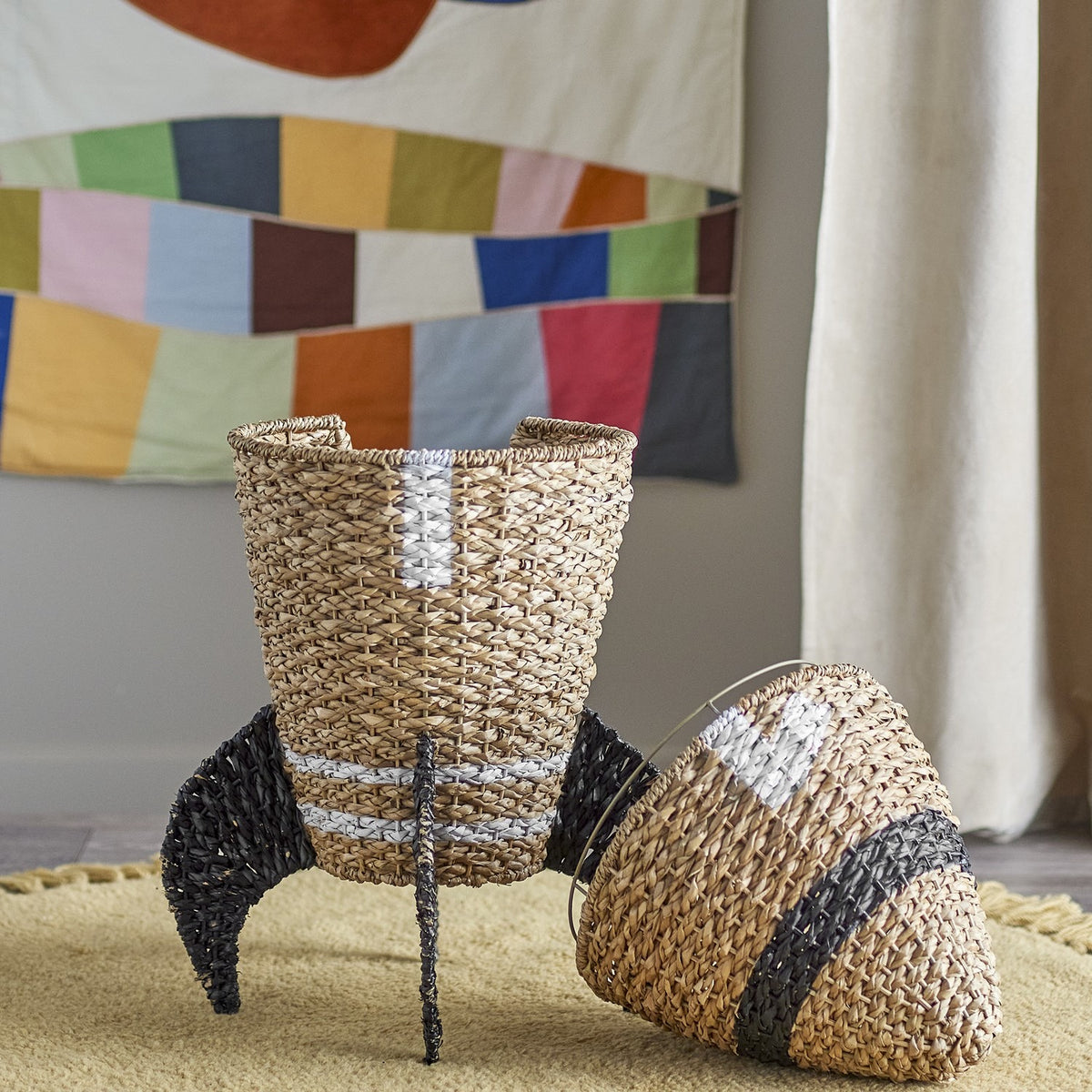 Cosmina Rocket Seagrass Basket with Lid in natural weave, shown as a rocket-shaped storage basket with black fin details and a cone-shaped lid beside it, set on a rug indoors.