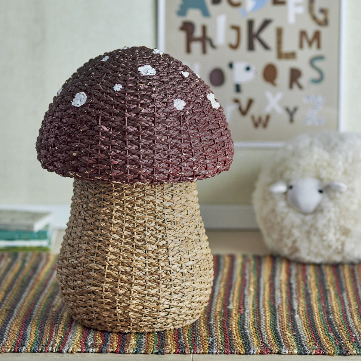 Corintha Mushroom Seagrass Basket with Lid in brown, domed spotted cap and woven stem, shown as nursery storage beside a plush sheep and alphabet print.