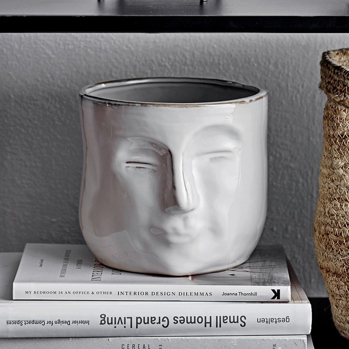 Ignacia Stoneware Plant Pot in white ceramic with a sculpted face in relief, closed eyes and full lips, shown on stacked books with a woven basket nearby.
