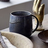 Cloe Speckled Stoneware Mug in dark blue-grey with handle, glaze streaking from rim into a speckled lower body, shown on a wooden table beside decorative dishes.