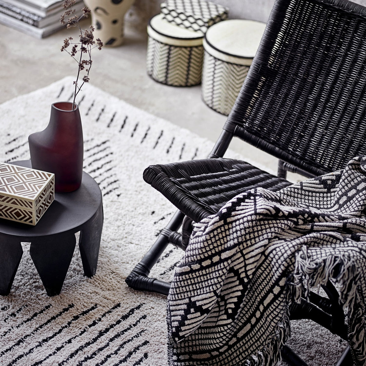 Gutte Geometric Throw in black and white draped over a woven black rattan chair, showing its diamond pattern, checks and fringed edge in a living room setting.
