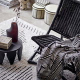 Gutte Geometric Throw in black and white draped over a woven black rattan chair, showing its diamond pattern, checks and fringed edge in a living room setting.