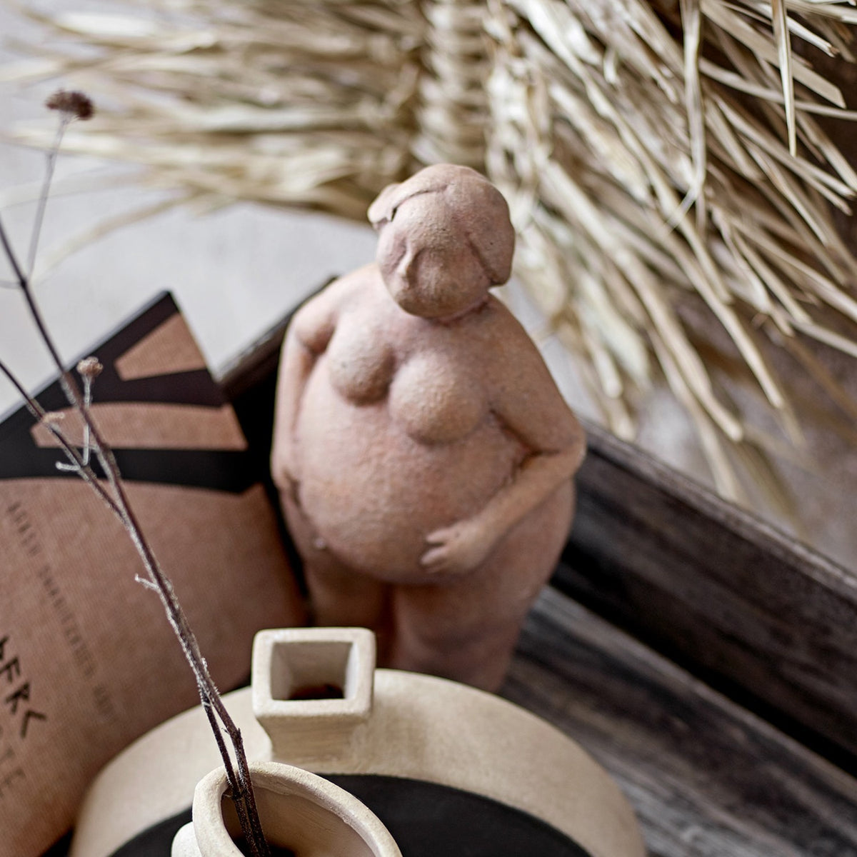 Sidsel Terracotta Ornament in brown, a standing female figurine with rounded curves and hands resting on her belly, displayed among dried grasses and decor.