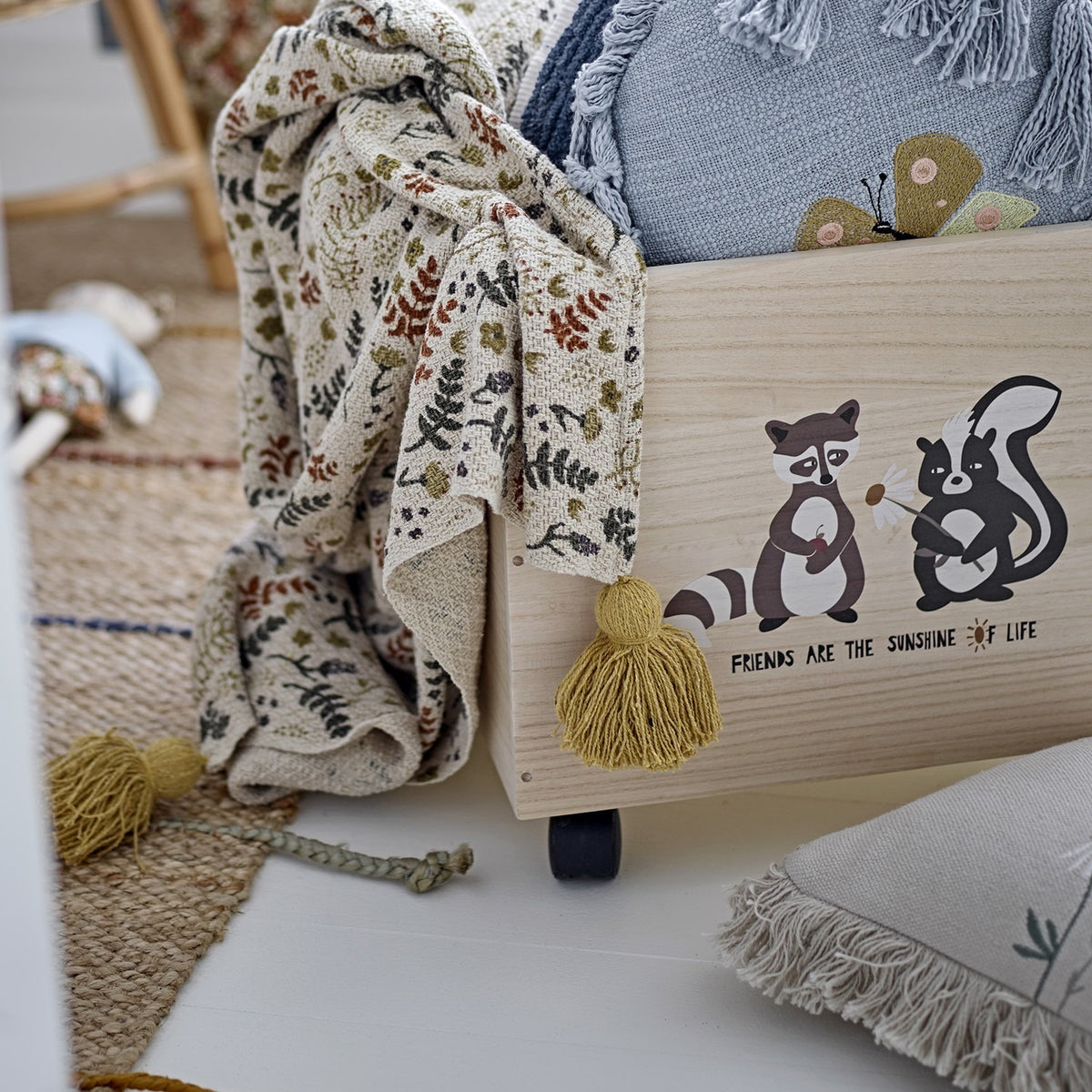 Filipa Floral Cotton Throw in multicoloured fern and leaf print with mustard tassel corners, draped over a toy storage box in a nursery setting.