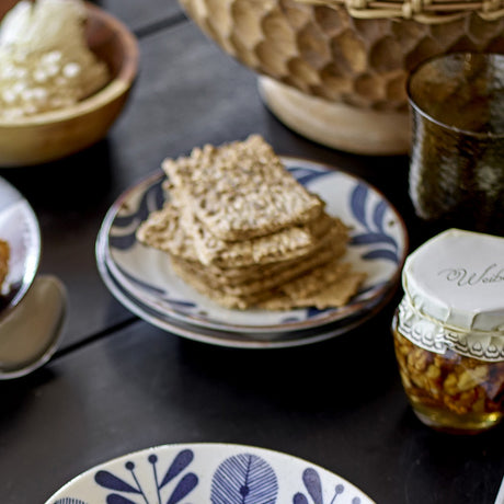 Camellia Porcelain Plate in cream with blue leaf border, holding stacked crackers on a dining table beside bowls, a jar, and a glass.
