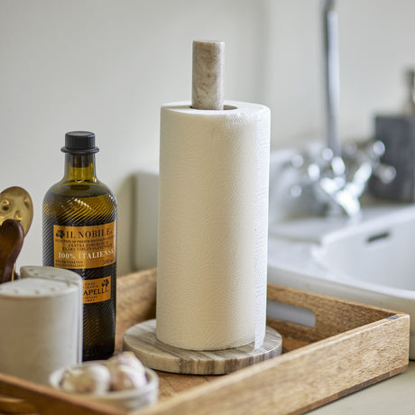 Emy Marble Kitchen Roll Holder in beige with natural veining, a slim round post on a flat base holding a paper towel roll, styled on a wooden tray beside oil and garlic.