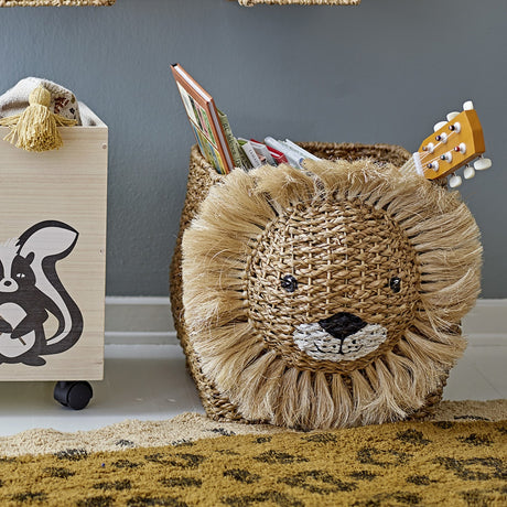 Harrison Basket in beige woven bankuan grass with a fringed lion's-head front, shown in a nursery filled with books and a toy guitar.