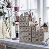 Richie Wooden Advent Calendar in natural plywood with 24 numbered drawers beneath a tiered winter village of houses, trees and deer, with built-in light.