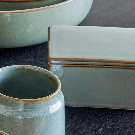 Pixie Stoneware Butter Dish in green, rectangular with a flat lid and brown rim edge, shown beside matching bowls and a mug in a reactive glazed finish.