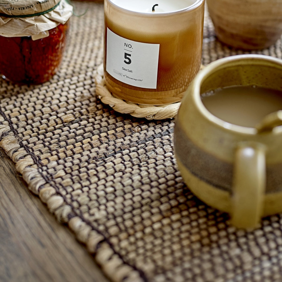 Solange Stoneware Mug 350ml in green, rounded stoneware body with loop handle, filled with coffee, shown on a woven jute table runner beside a candle and jar.