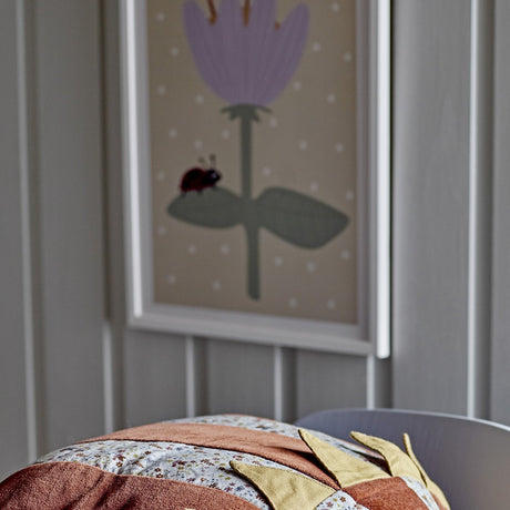 Numa Wooden Framed Print in multi, shown on a nursery wall above a patterned cushion, with a pale purple flower, green leaves and a ladybird on a spotted beige background.