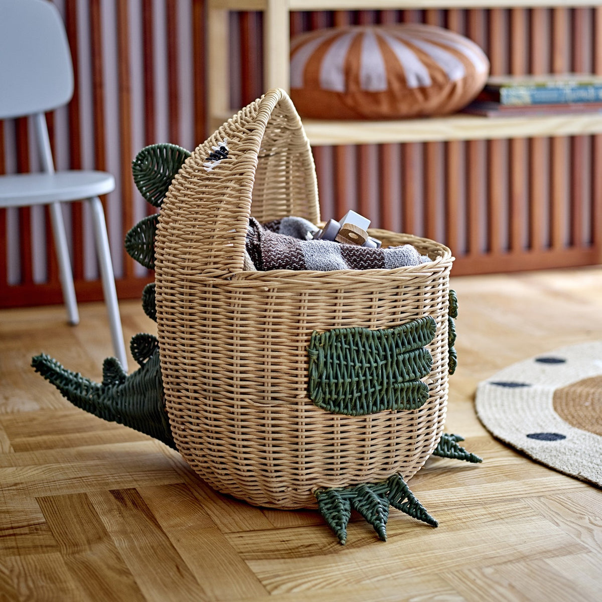 Eddi Dinosaur Rattan Basket in natural with green woven spines, arms and clawed feet, shown holding blankets on a wooden floor in a living room setting.