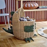 Eddi Dinosaur Rattan Basket in natural with green woven spines, arms and clawed feet, shown holding blankets on a wooden floor in a living room setting.
